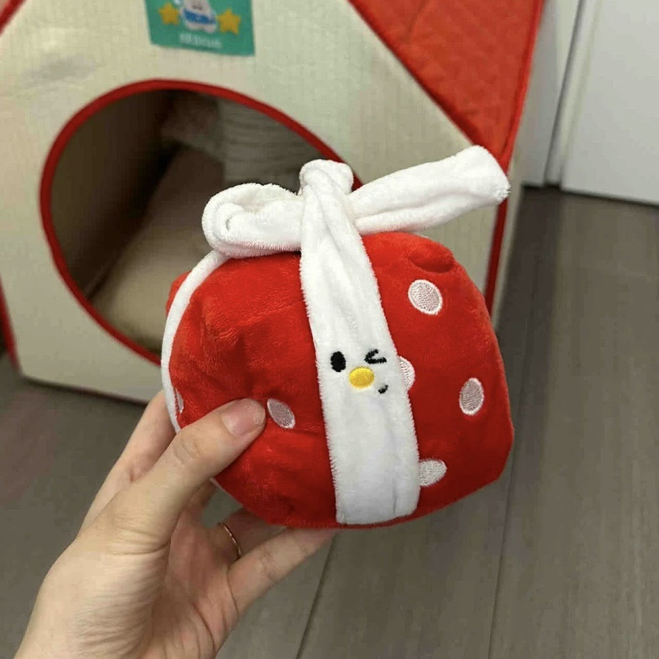 Red plush toy with a white bow held by a hand, with a pet bed in the background.