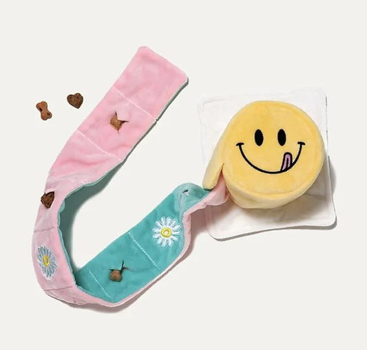 A colorful dog nosework toy designed to look like a smiley cake with pockets of cloth for hiding treats, laid flat with treats scattered around.