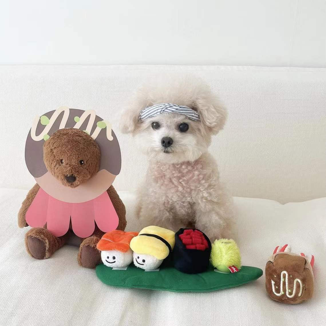 Small dog sitting next to a set of plush toys shaped like a bear, sushi rolls, and a leaf on a white background.