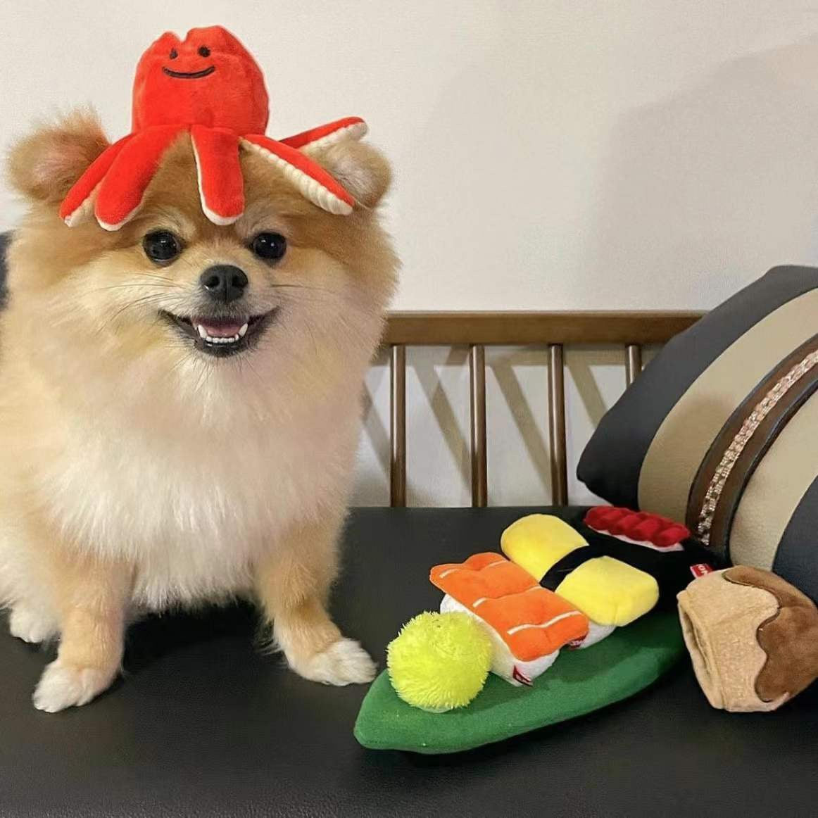 Dog wearing a crab hat with a toy resembling sushi on a bed